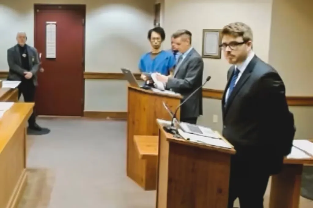 Makoto Kuroda in court, wearing a blue shirt, stands beside an attorney while facing the judge.