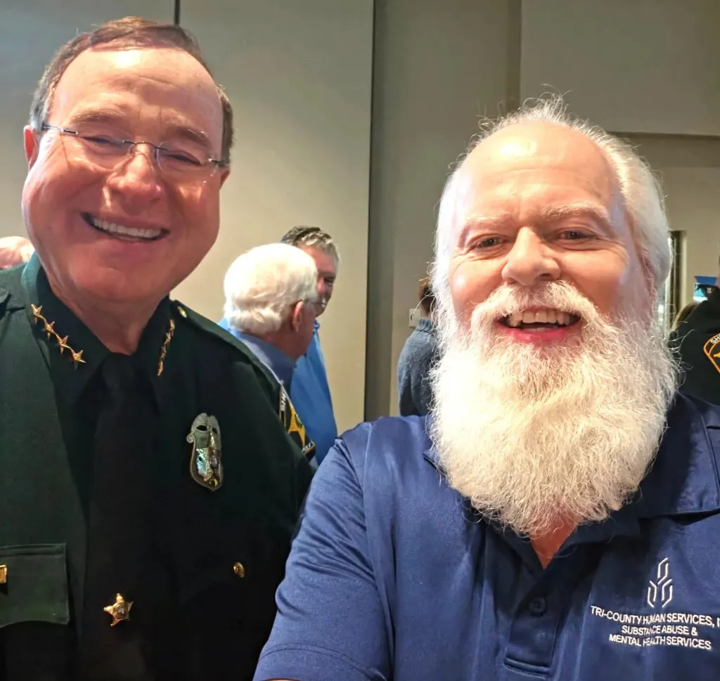 Thomas Hicks, wearing a blue shirt with "TRI-COUNTY HUMAN SERVICES" on it, stands next to a man in a police uniform.