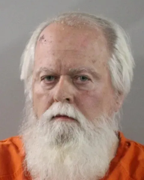 Mugshot of Thomas Hicks, a man with white hair and a long white beard, wearing an orange jumpsuit.