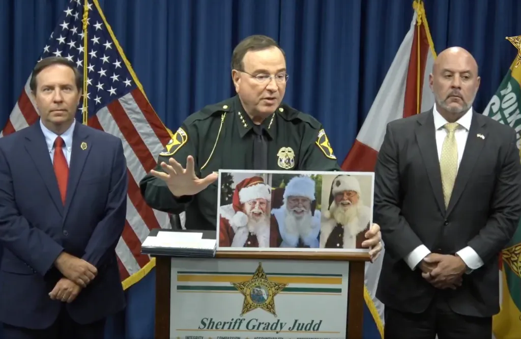 Sheriff Grady Judd speaking at a press conference, holding up a picture of three men dressed as Santa Claus.