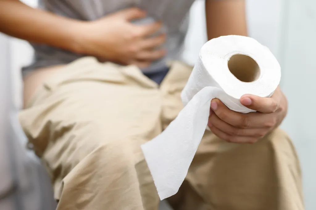 A person sitting on a toilet with their pants down, holding a roll of toilet paper, and clutching their stomach.
