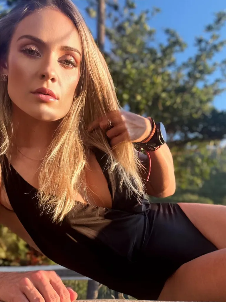 A woman in a black swimsuit with blonde hair and light makeup poses for a photo, with trees and blue sky in the background.