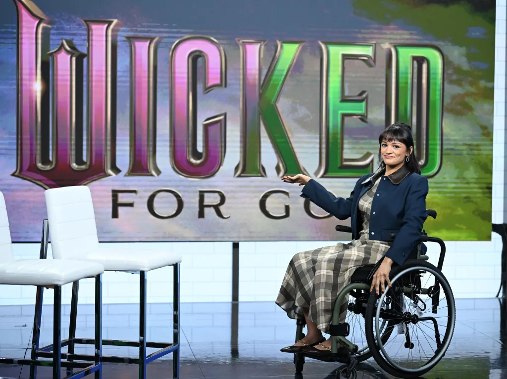 Marissa Bode in a wheelchair, gesturing with one hand in front of a "Wicked for Good" sign.