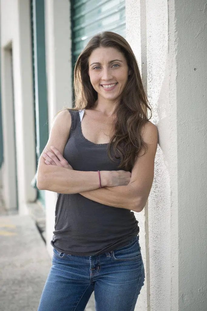 Mary Padian smiling with her arms crossed, wearing a dark gray tank top and blue jeans, leaning against a white pillar.