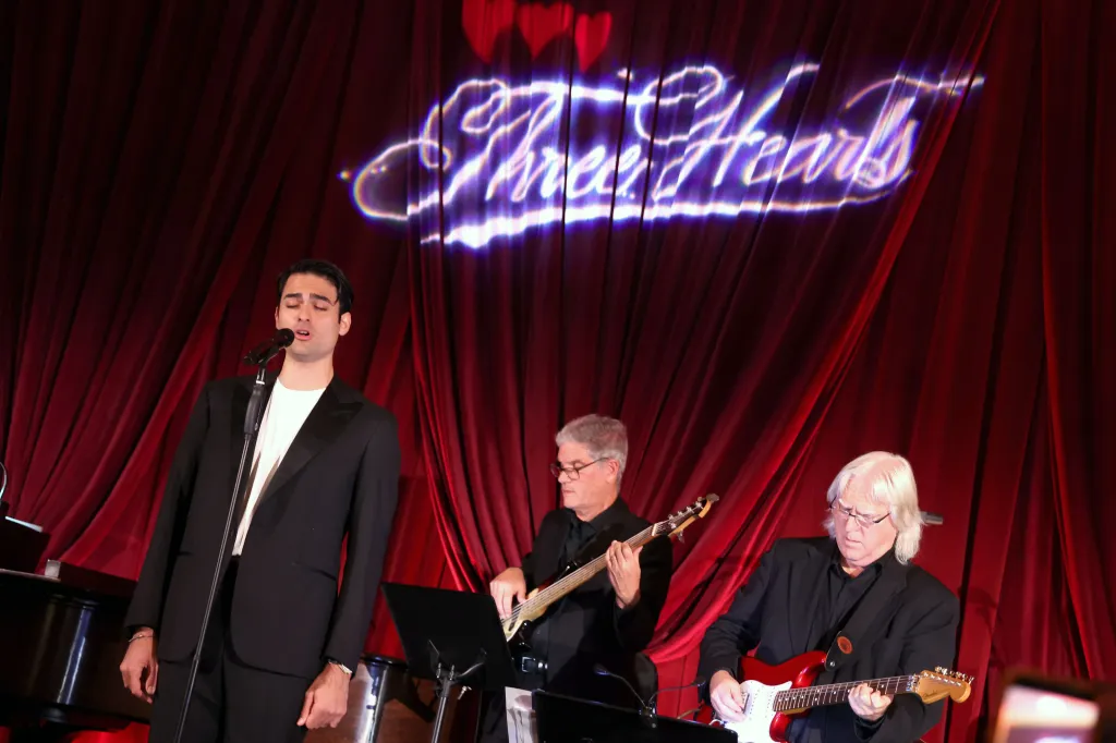 Matteo Bocelli sings into a microphone with two guitarists on stage, in front of a red curtain with the words "Three Hearts" projected onto it.