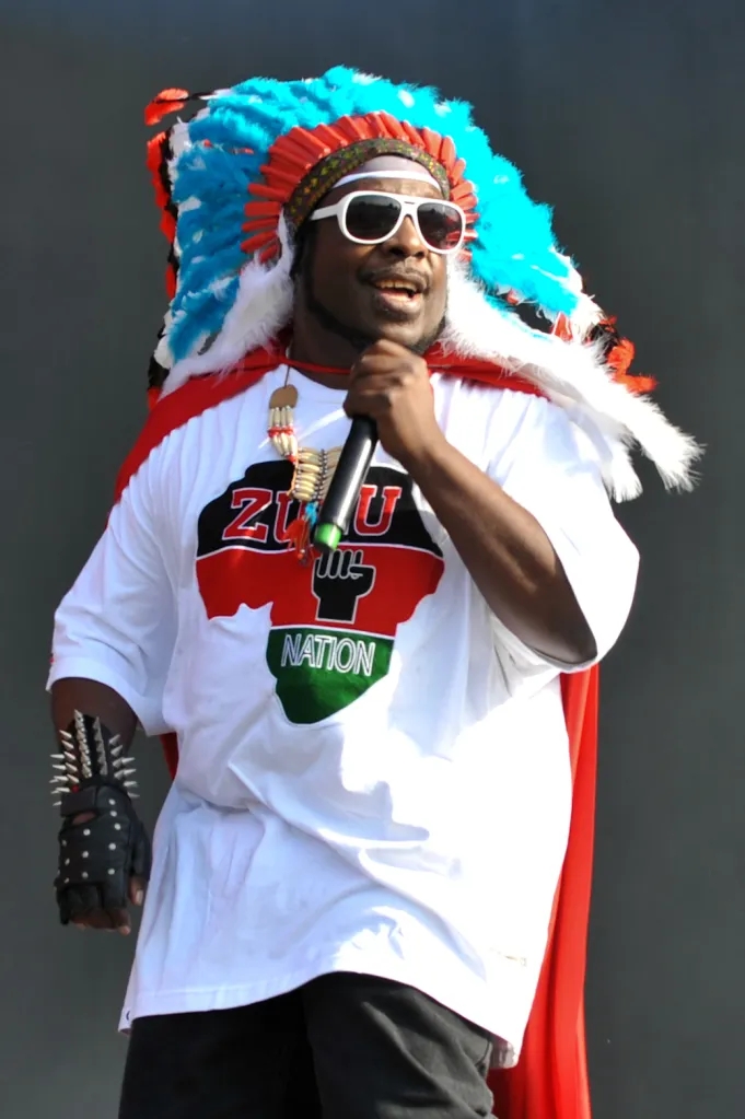 A man wearing a feathered headdress, sunglasses, and a "Zulu Nation" t-shirt, holding a microphone.