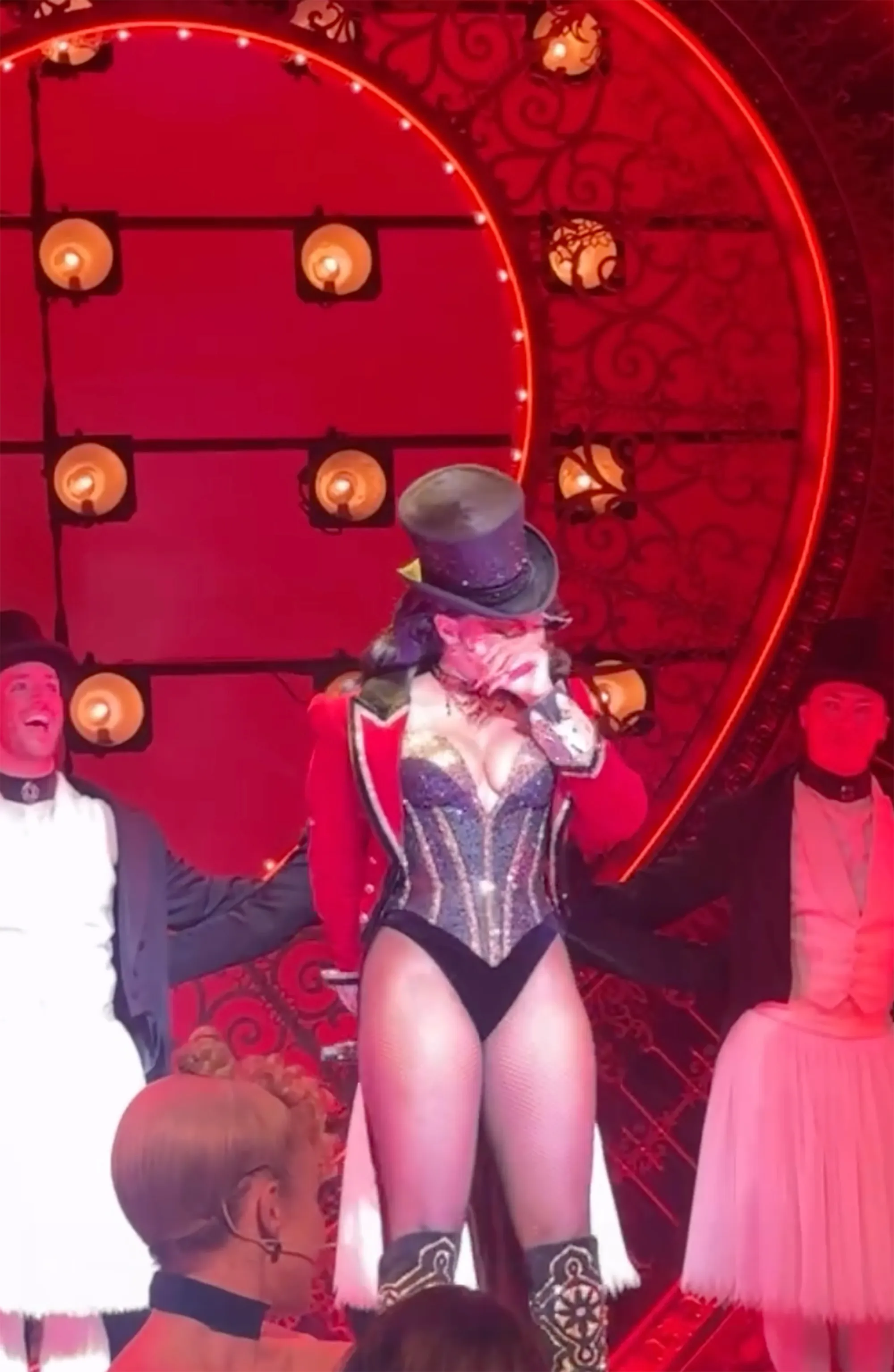 Megan Thee Stallion in a black corset and red jacket, wearing a top hat and wiping tears from her face on stage during a Broadway performance.