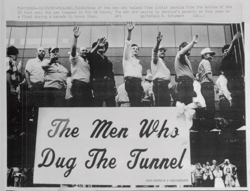 Men who rescued Jessica McClure wave during a parade, behind a banner that reads "The Men Who Dug The Tunnel."