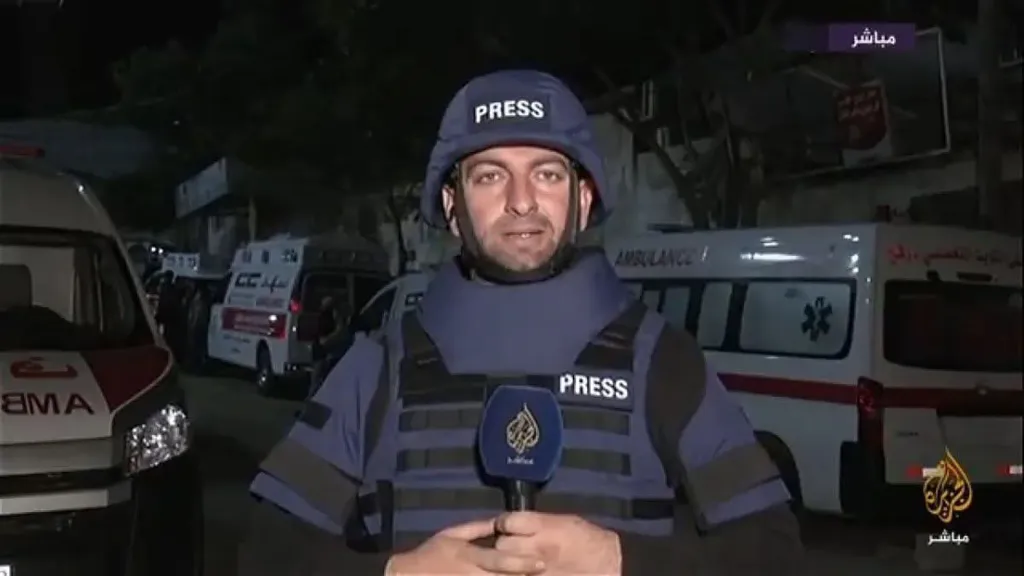 Muhammad Samir Muhammad Washah, an Al Jazeera journalist wearing a "PRESS" helmet and bulletproof vest, reporting in front of ambulances.