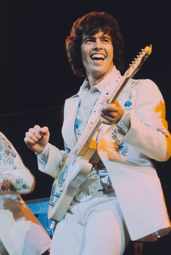 Alan Osmond performs live on stage, playing a guitar and wearing a white suit with blue and green floral embroidery.