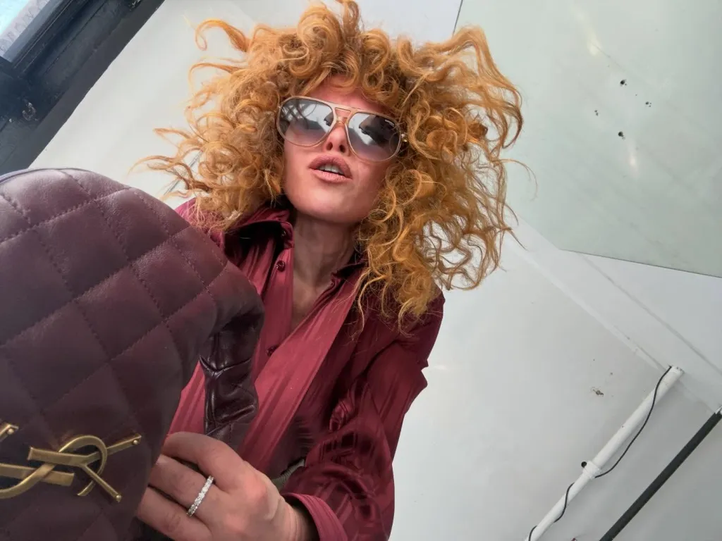 Natasha Lyonne in a selfie, wearing aviator sunglasses and holding a quilted burgundy handbag.