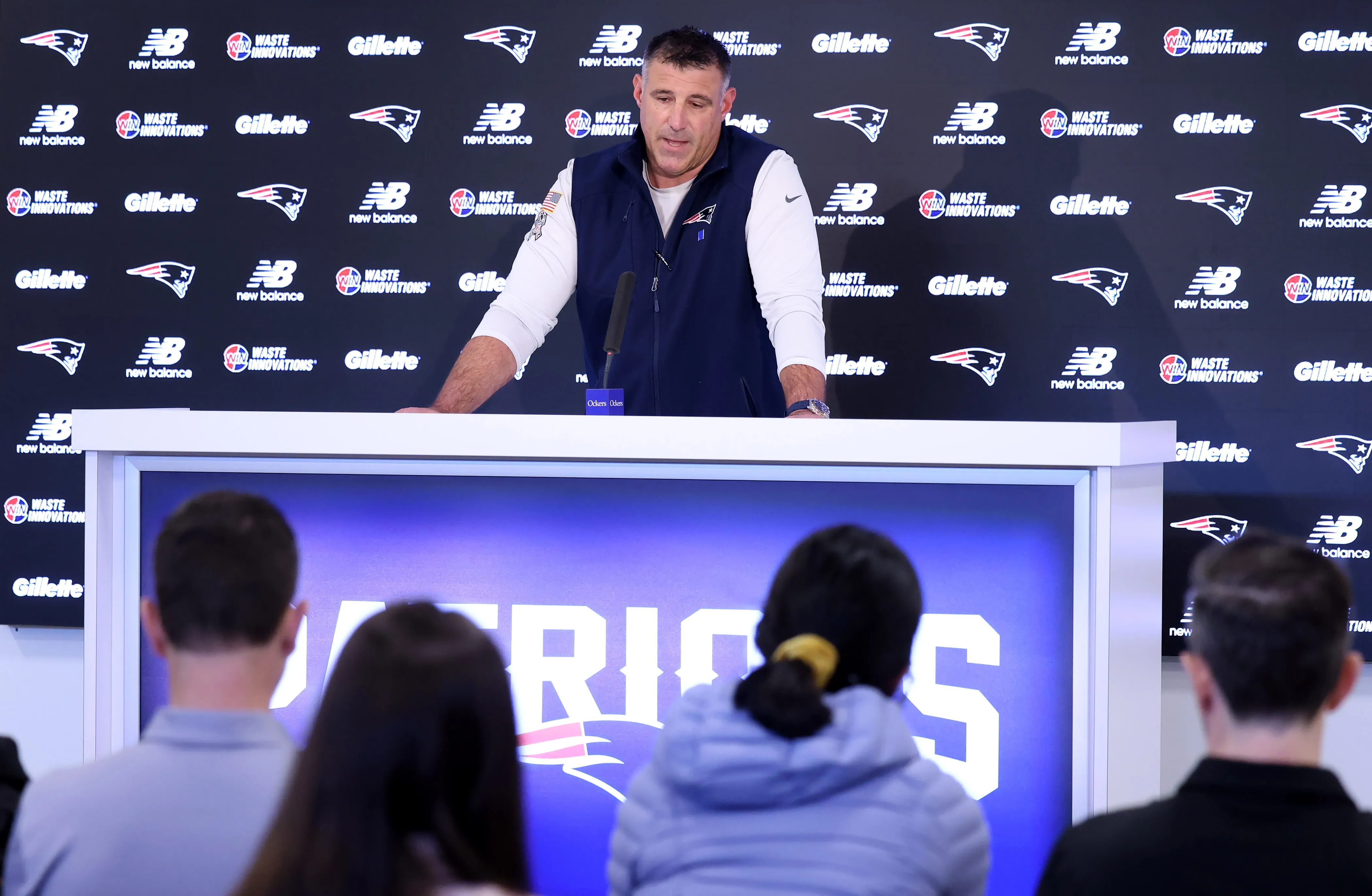 Mike Vrabel speaking at a press conference with the New England Patriots logo visible.