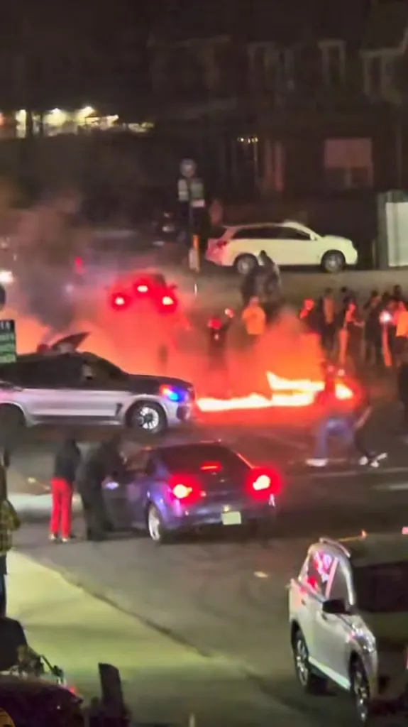 A street at night with cars and a crowd of people gathered around a large fire.