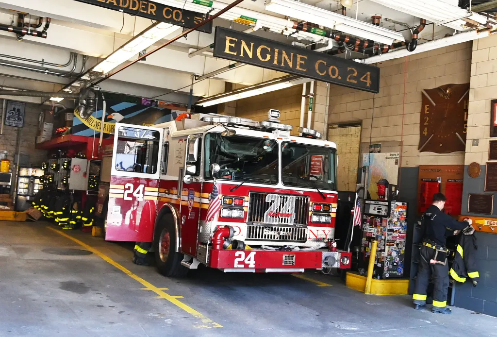 A red and white FDNY Engine 24 fire truck sits inside its firehouse as a firefighter prepares his gear.
