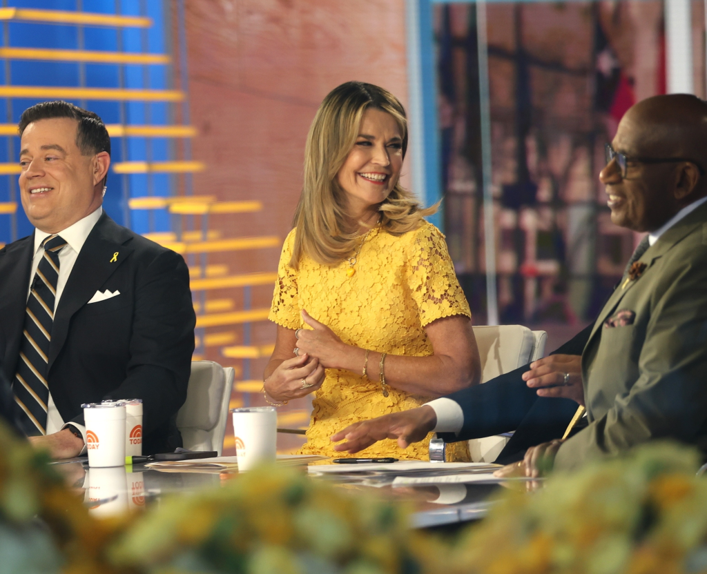 Savannah Guthrie, wearing a yellow dress, sits between her "Today" show co-hosts, Craig Melvin and Al Roker, at the anchor desk.
