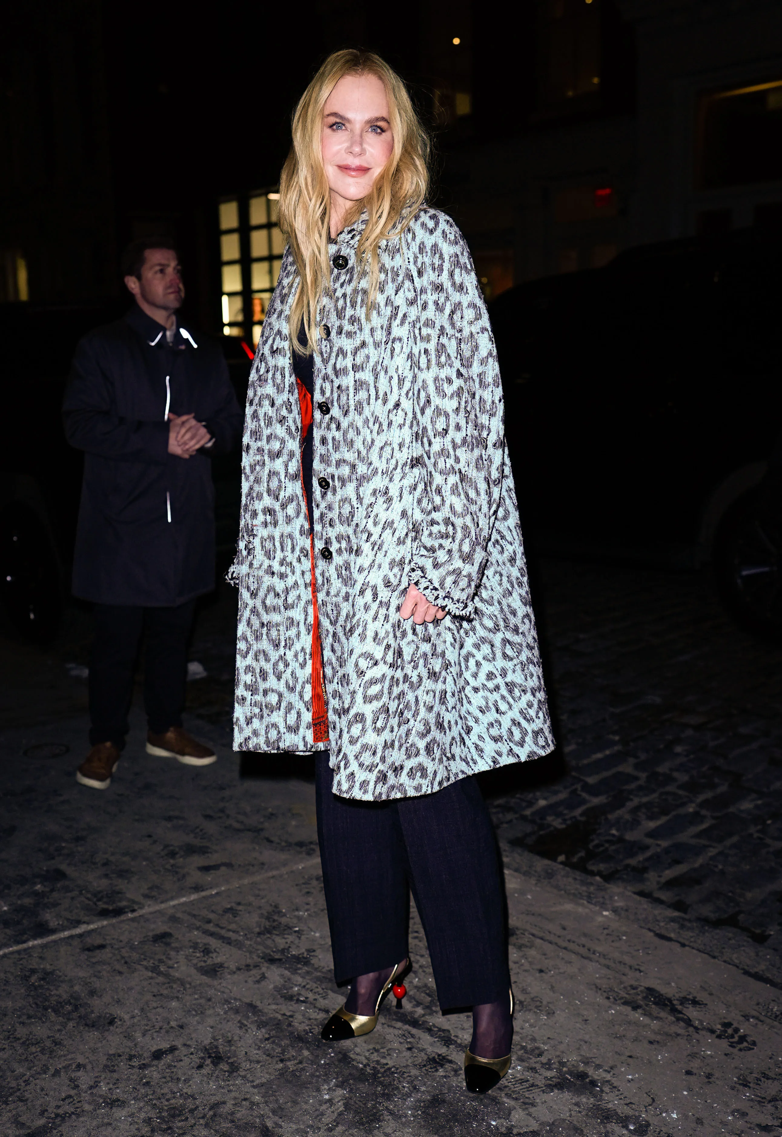 Nicole Kidman standing outside wearing a blue and black leopard print coat and black pants.