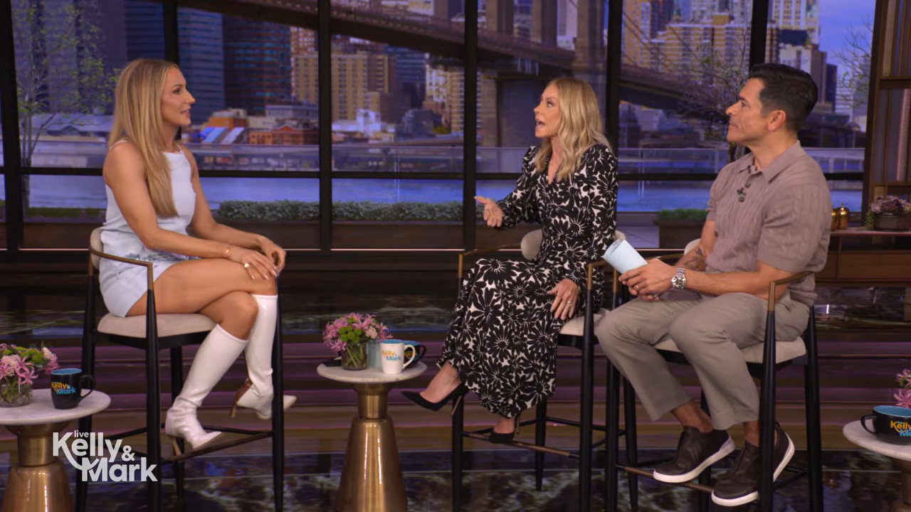 Nikki Glaser in a light blue mini dress, Kelly Ripa in a black and white floral dress, and Mark Consuelos in a grey polo shirt sit on a stage with a city skyline backdrop.