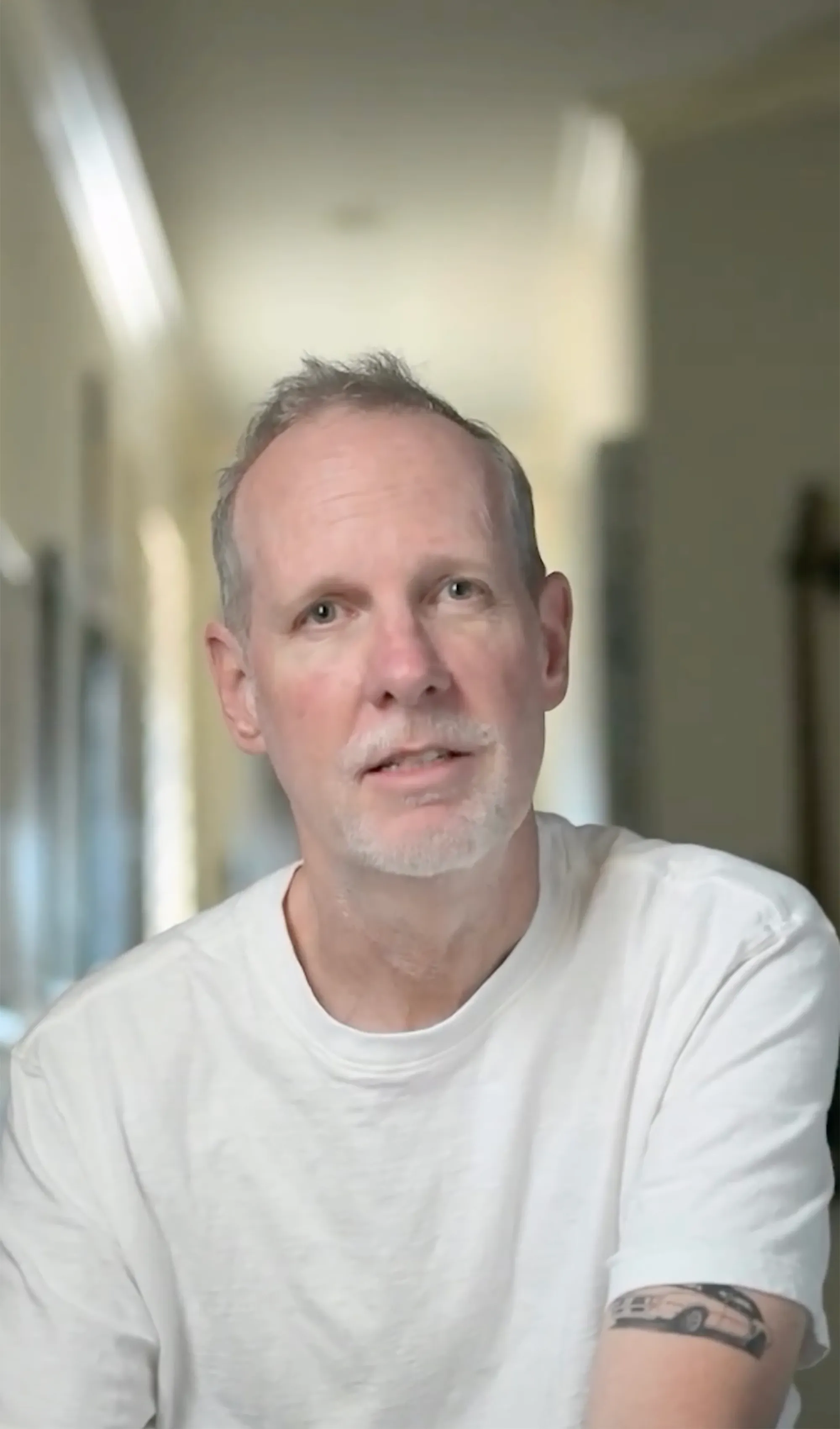 Tom Dumont, guitarist for No Doubt, shown from the chest up, wearing a white t-shirt with a tattoo of a white car on his left arm.