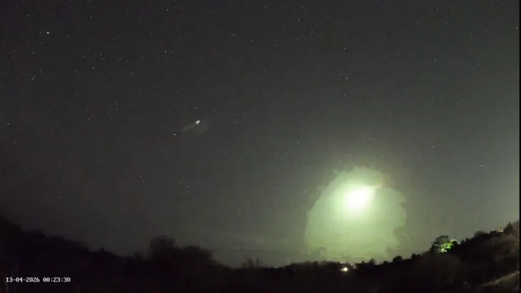A green fireball streaks across a starry night sky above a dark landscape with some illuminated structures.