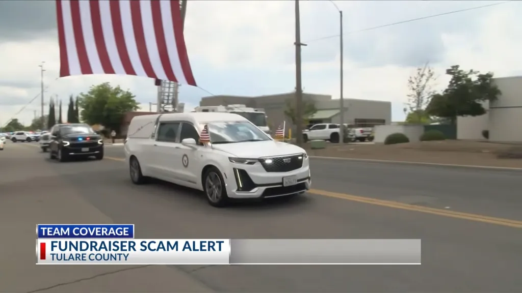 A white hearse leads a procession of vehicles down a road with a large American flag draped overhead.