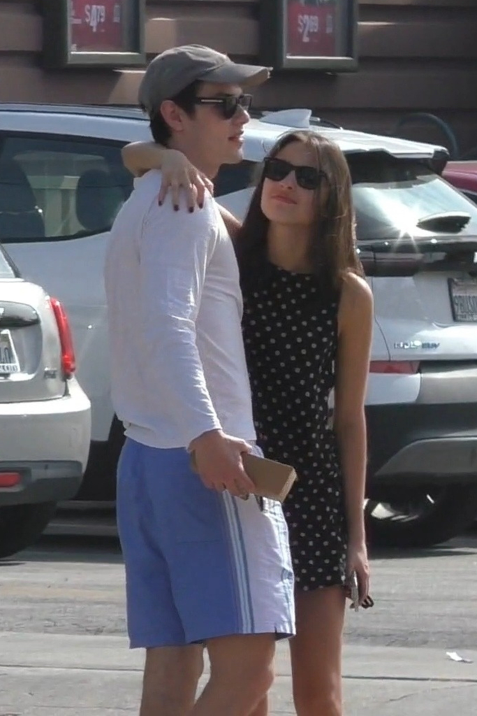 Olivia Rodrigo, wearing sunglasses and a polka-dot dress, and Louis Partridge, wearing a cap and sunglasses, embrace after lunch.
