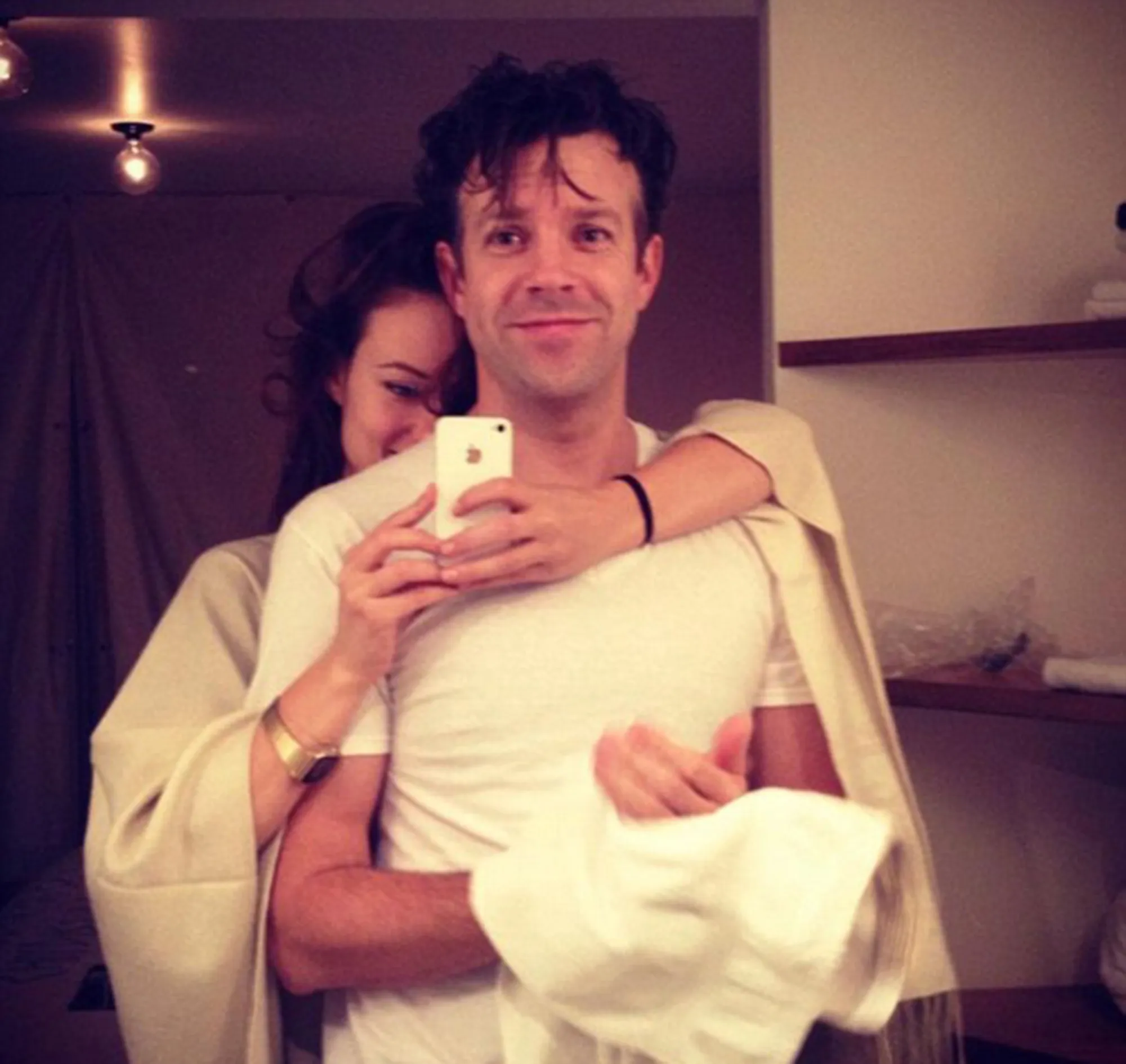 Olivia Wilde smiles behind Jason Sudeikis as he takes a mirror selfie.