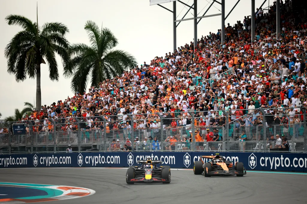 Max Verstappen leading Lando Norris in a Formula 1 race at Miami International Autodrome, with a crowd of spectators in the background.