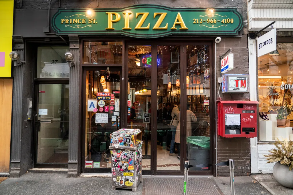 An exterior view of Prince St Pizza, with a green sign over the door and an "Out of Order" ATM to the right.