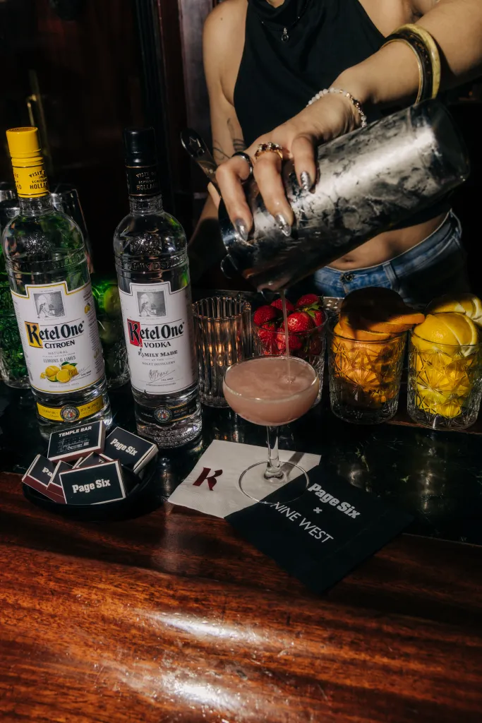 A bartender pours a drink into a cocktail glass, surrounded by Ketel One vodka bottles, Page Six x Nine West branded items, and fruit.