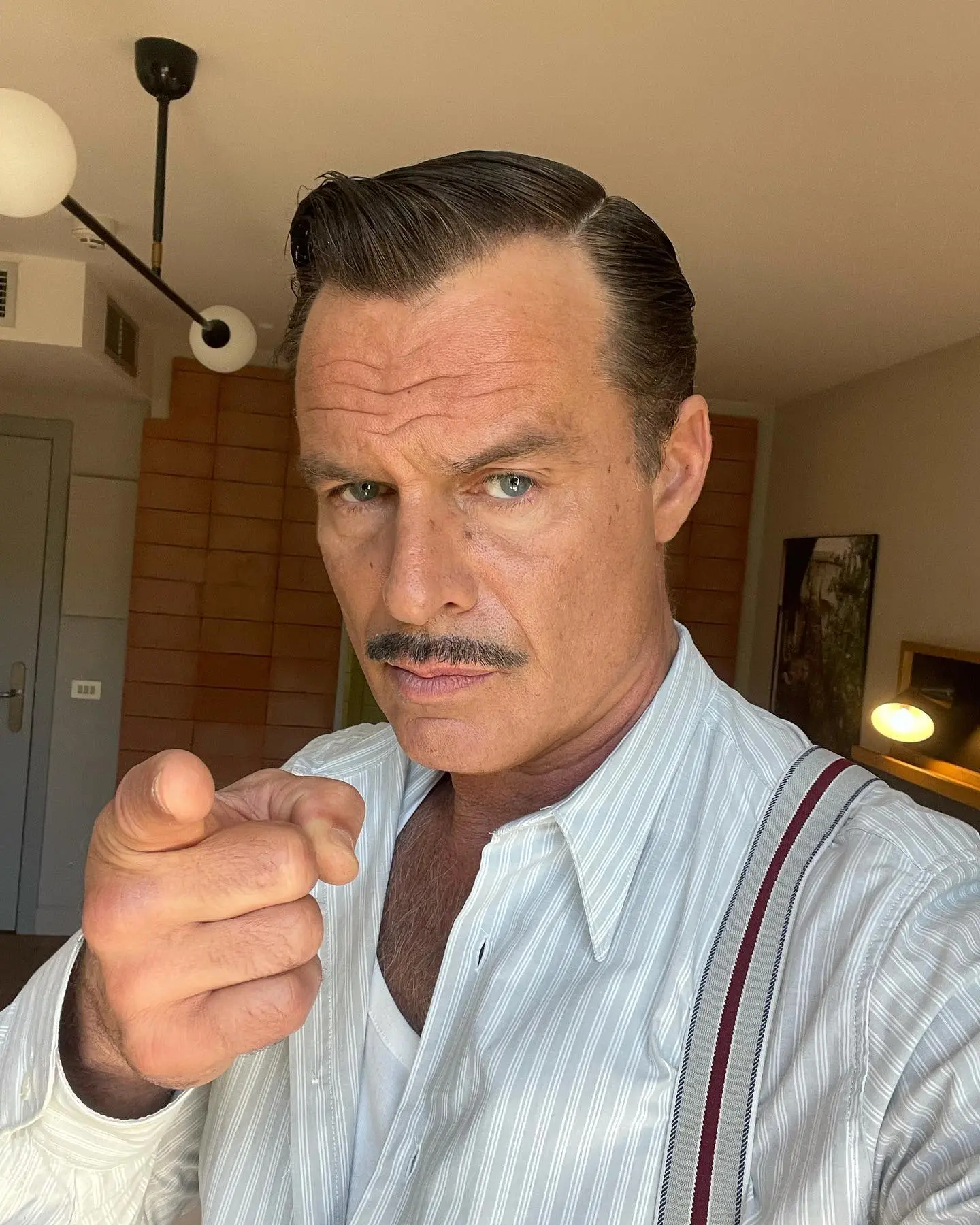 Patrick Muldoon posing for the camera, pointing at the viewer, with a mustache and wearing a pinstriped shirt and suspenders.