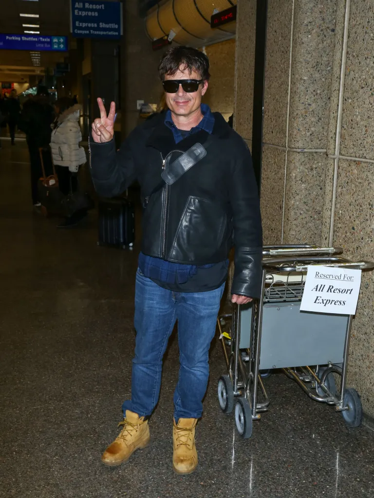 Patrick Muldoon wearing sunglasses and a black leather jacket, with jeans and work boots, gives a peace sign at Salt Lake International Airport.