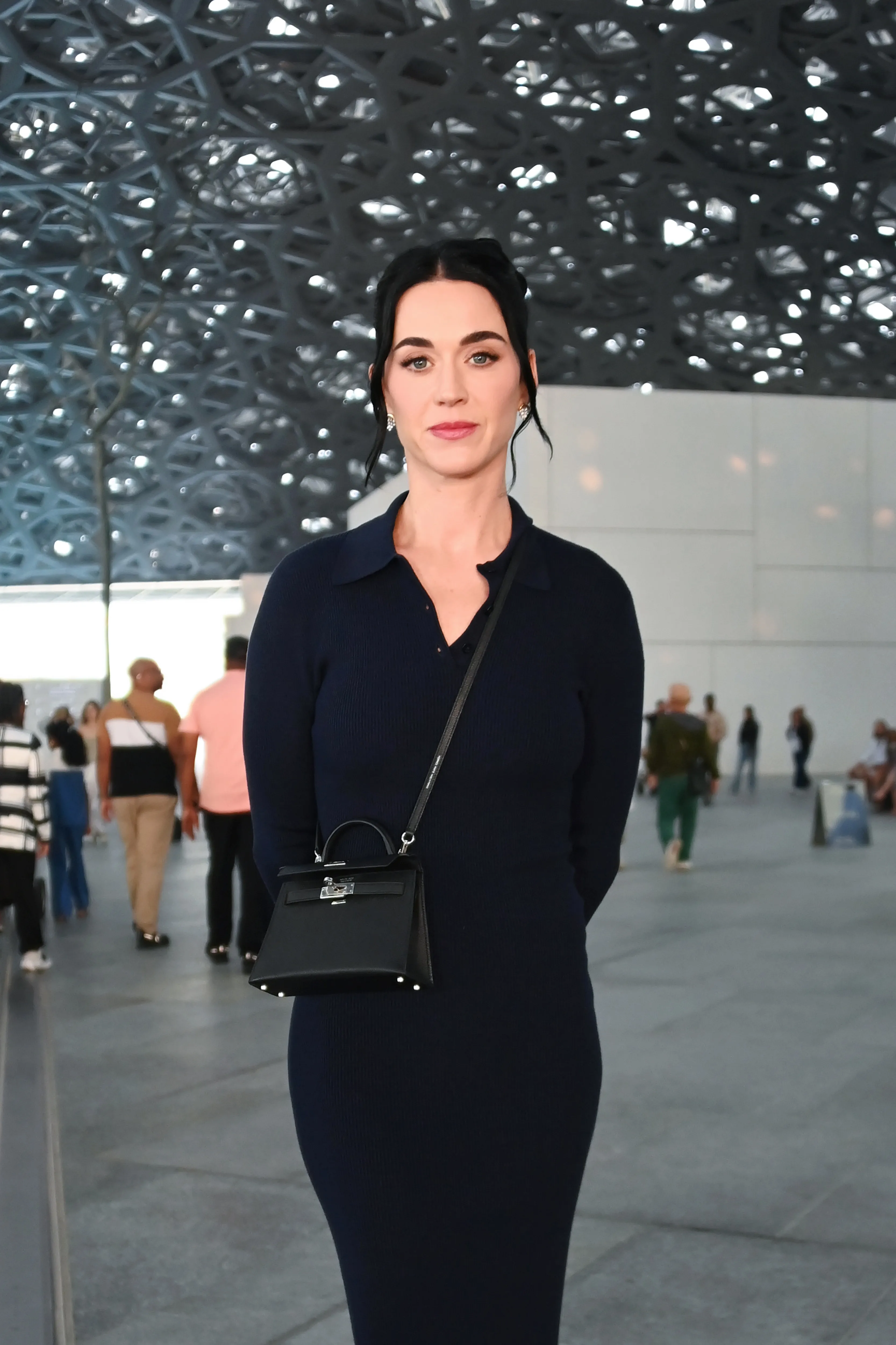 Katy Perry poses in a dark blue collared dress and black handbag with the Louvre Abu Dhabi's patterned dome roof in the background.
