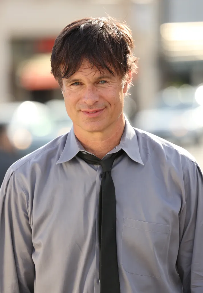 Patrick Muldoon poses wearing a light gray shirt and black tie.