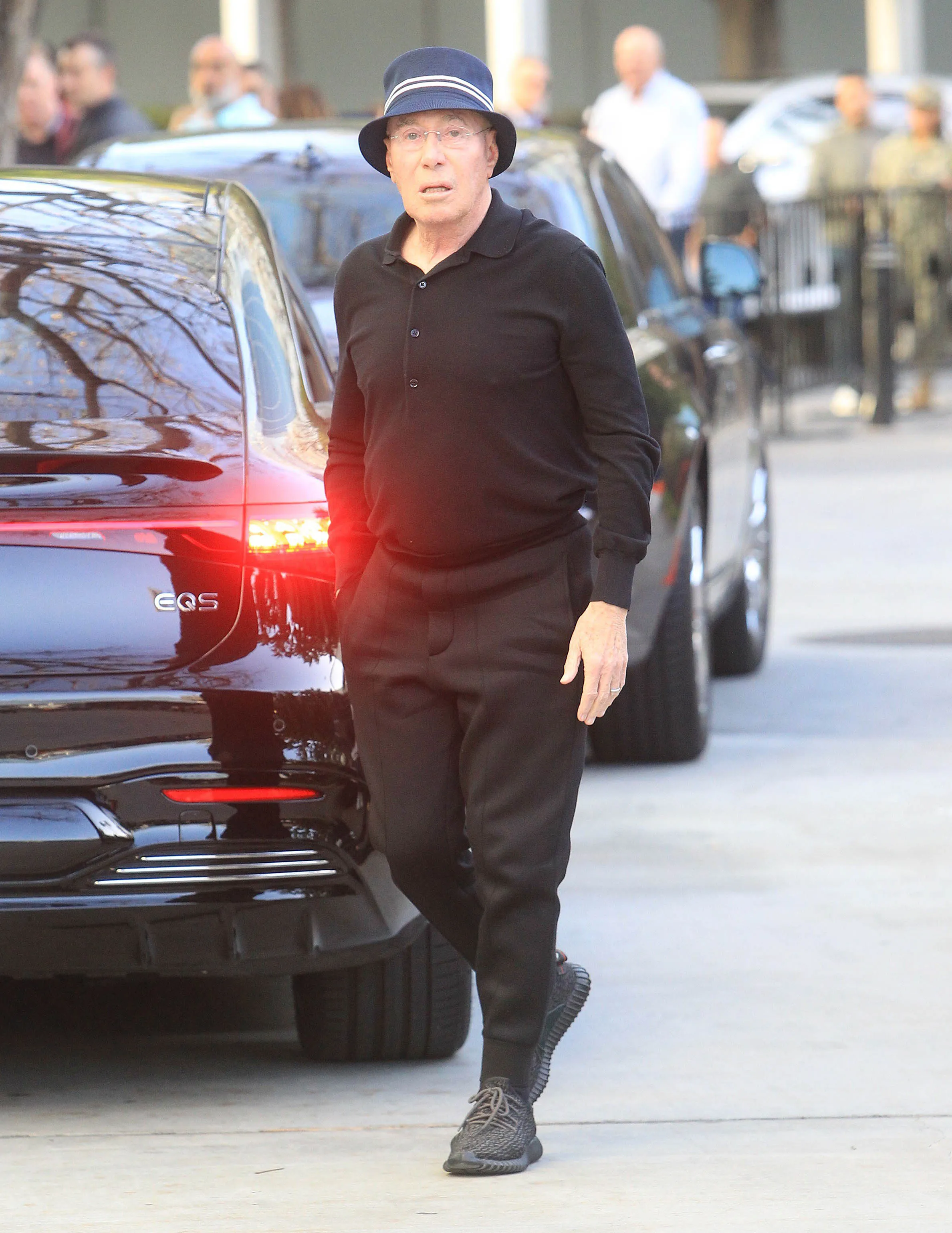 David Geffen walking outdoors in a blue hat, black polo shirt, and black pants, next to a black car.