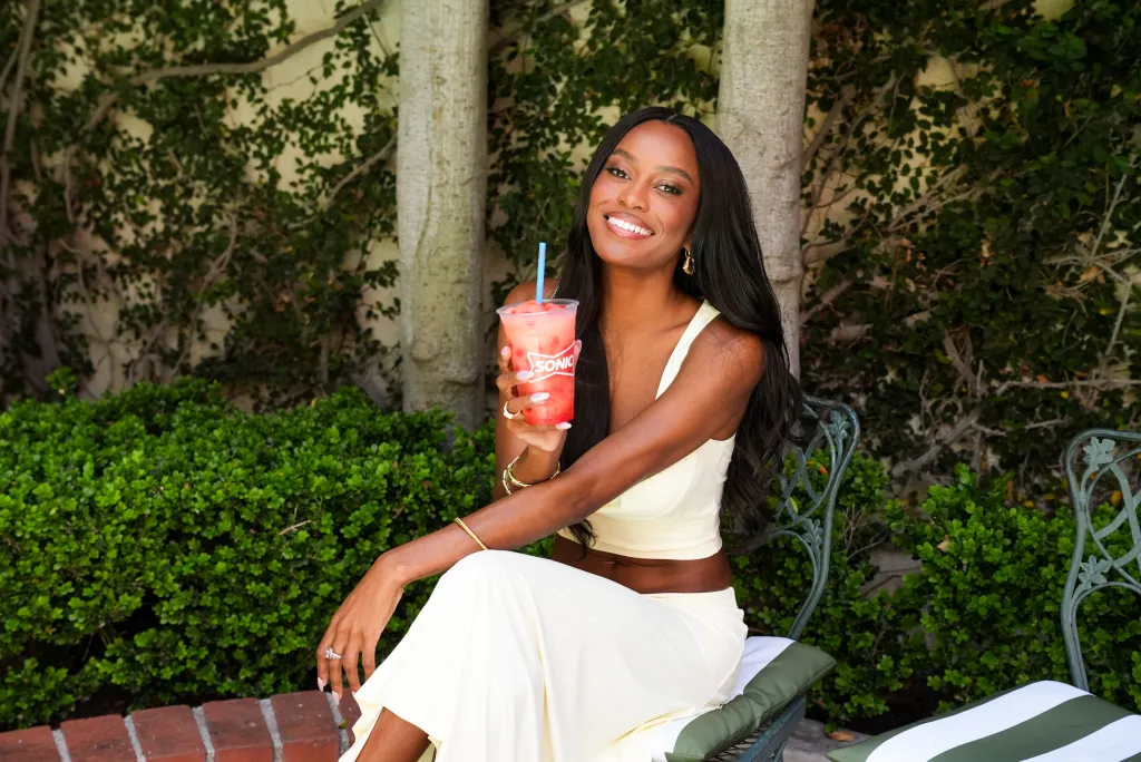 Ciara Miller sits on a chair, smiling at the camera and holding a Sonic Refresher drink.