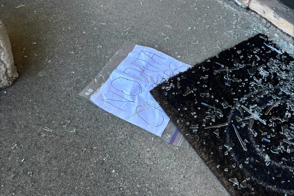 Shattered glass on a black doormat and ground, with a "NO" sign in a plastic bag next to it.