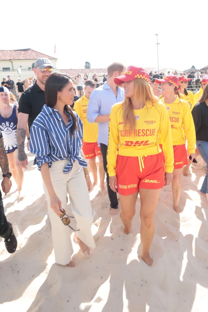 Meghan Markle and Prince Harry walking barefoot on Bondi Beach with lifeguards.