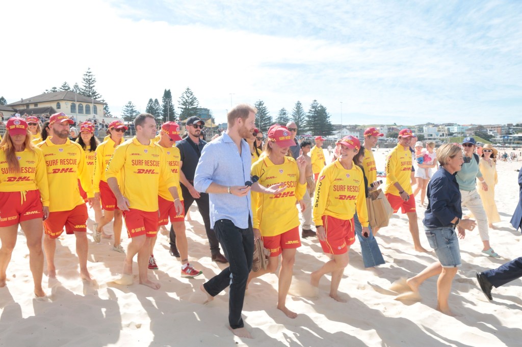Prince Harry walks barefoot on Bondi Beach with lifeguards in yellow and red uniforms.