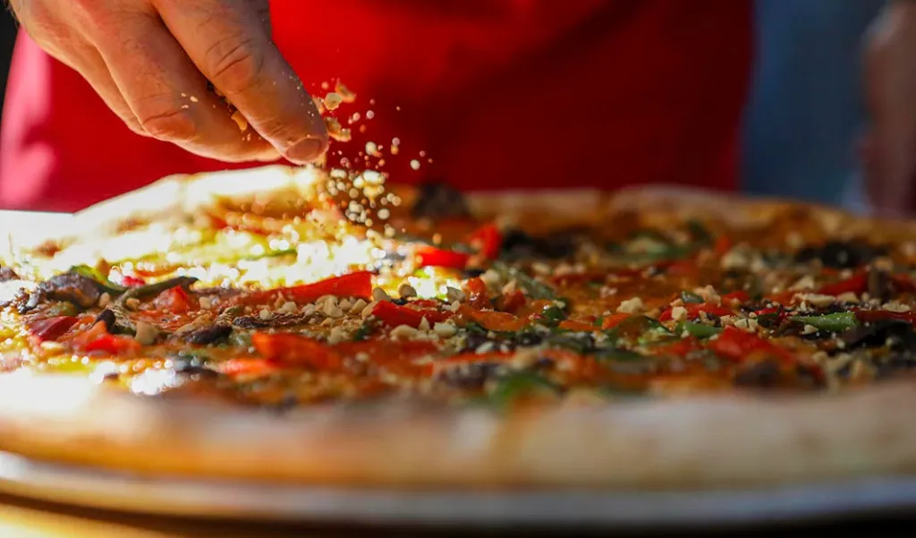 A hand sprinkles toppings over a pizza with red peppers and green vegetables.