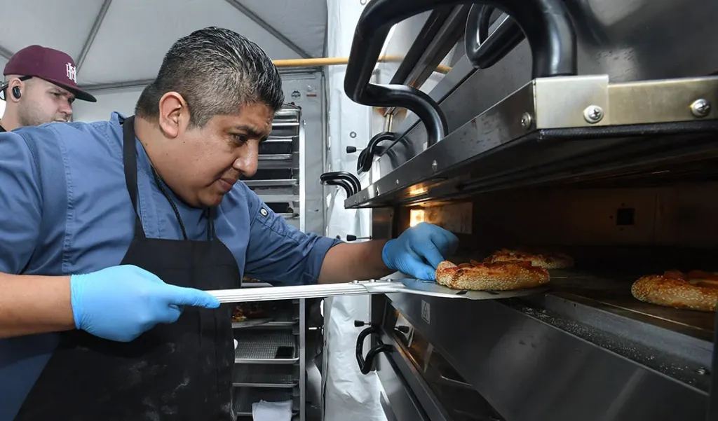 A chef in a blue shirt and black apron, wearing blue gloves, sliding a flatbread pizza into an oven.