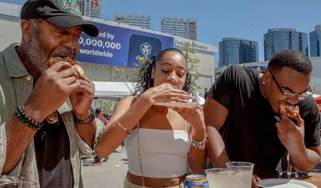 Three people eating pizza at an outdoor festival.