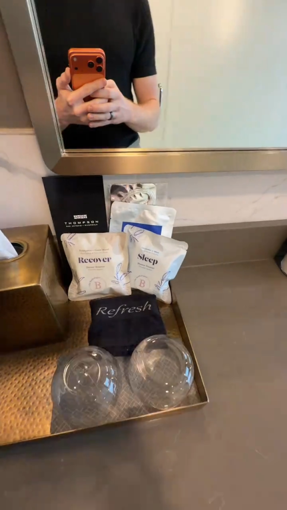 Hotel amenities like "recover" and "sleep" shower steamers, and "refresh" washcloth are on a tray next to two glasses, appearing to be free but are chargeable.