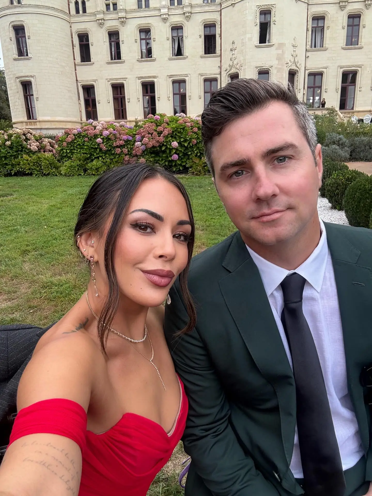 Janel Parrish and Chris Long pose for a selfie with a building and garden in the background.