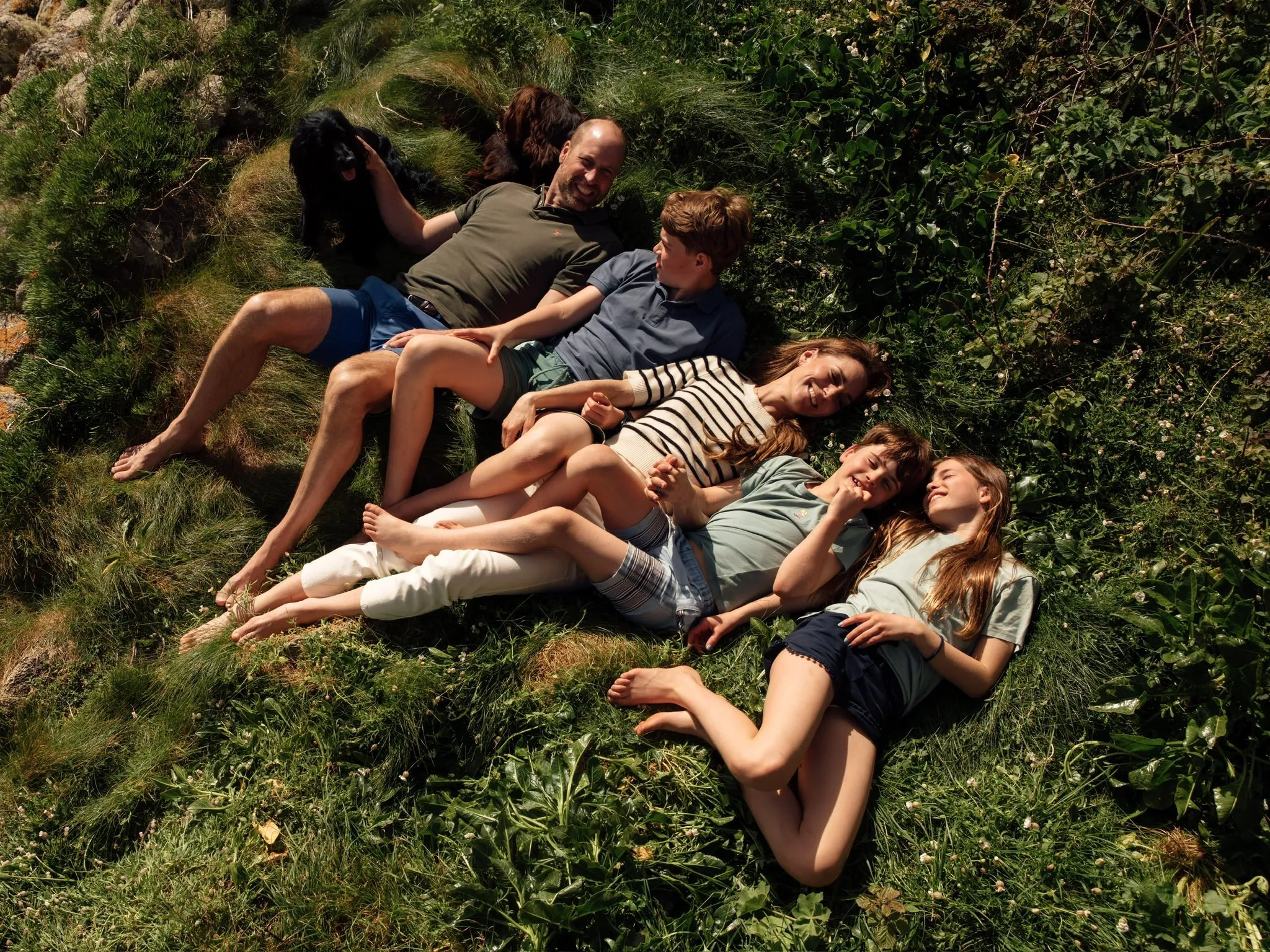 The Prince and Princess of Wales with their children and dog, lying on the grass.
