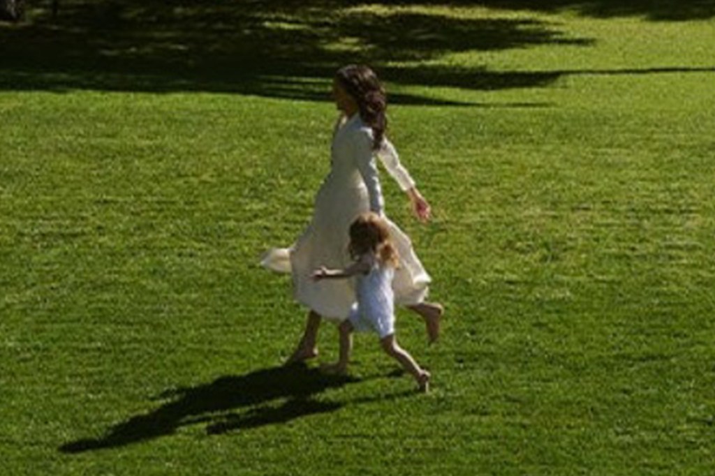 Meghan Markle, in a white dress, and Princess Lilibet, in a white romper, walking barefoot on a grassy field.