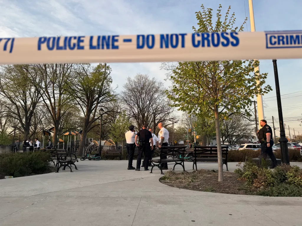 Police line "Do Not Cross" tape in the foreground of a park where several police officers are gathered.