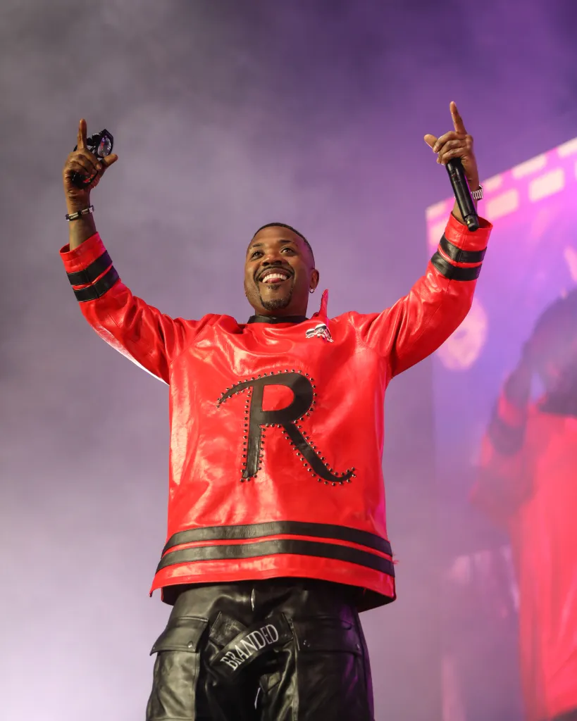 Ray J performing during The Millennium Tour, wearing a red jacket with a large "R" on it and holding a microphone and sunglasses.