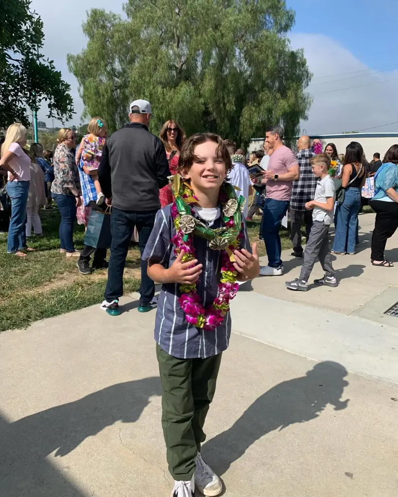 Reis Beck, a 13-year-old boy, smiles while wearing leis over his striped shirt and standing on a sidewalk with a crowd in the background.