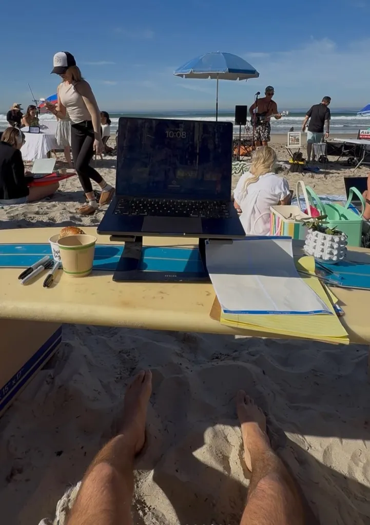 A remote work meetup on the beach with a laptop on a surfboard, a person's bare feet in the sand, and people networking in the background.