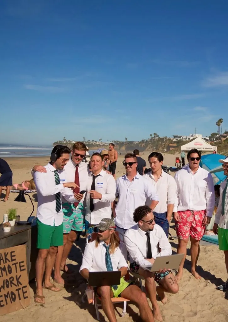 A group of men in shirts and ties with swim trunks at a "remote work meetup" on the beach.